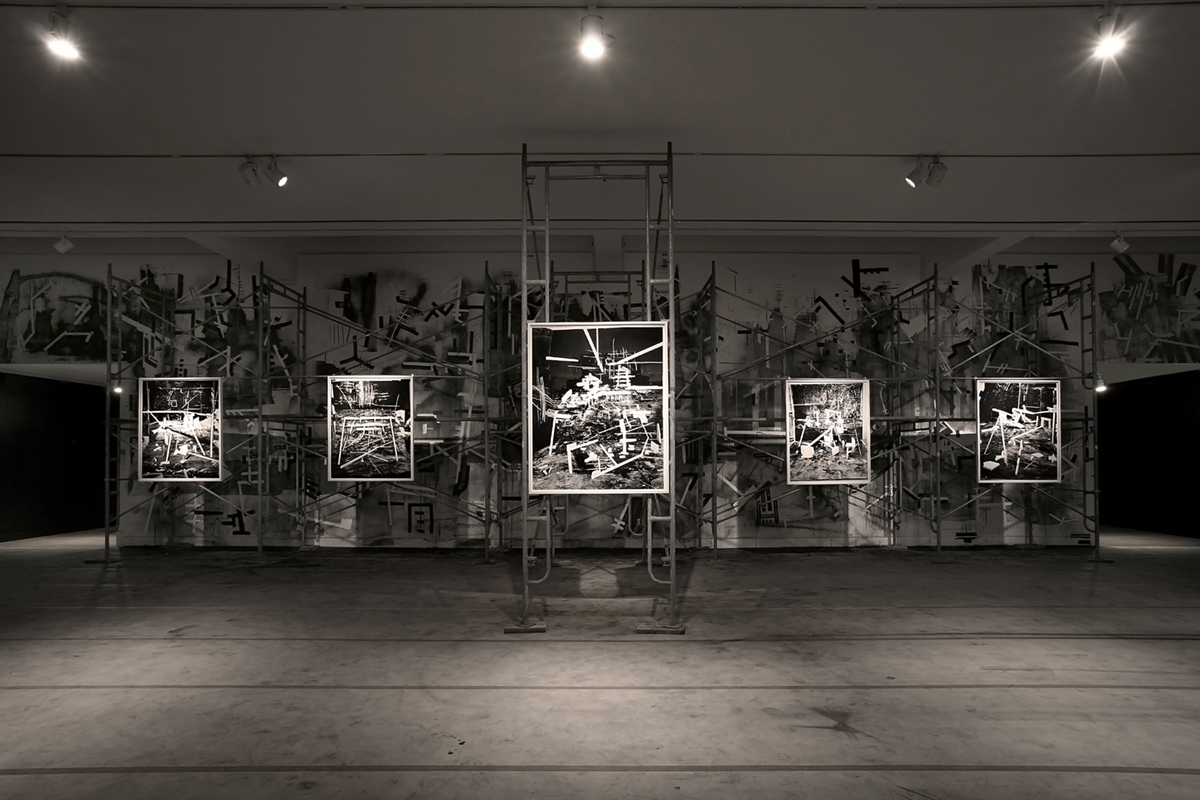 A dark gallery space showcases five black and white photographs that depict scenes of industrial materials and ruins in high contrast.  The photograph in the center hangs from scaffolding, and all the works are illuminated using spotlights. The back wall of the gallery is painted using black and white industrial motifs that echo the content of the photographs.