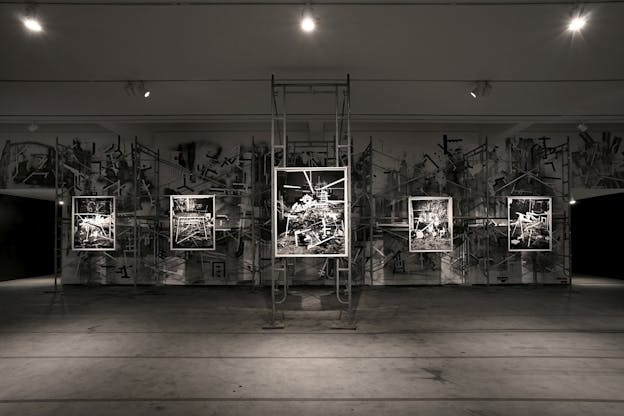 A dark gallery space showcases five black and white photographs that depict scenes of industrial materials and ruins in high contrast.  The photograph in the center hangs from scaffolding, and all the works are illuminated using spotlights. The back wall of the gallery is painted using black and white industrial motifs that echo the content of the photographs.