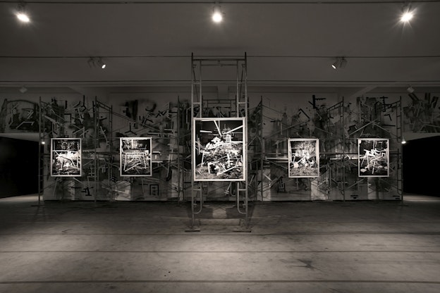 A dark gallery space showcases five black and white photographs that depict scenes of industrial materials and ruins in high contrast.  The photograph in the center hangs from scaffolding, and all the works are illuminated using spotlights. The back wall of the gallery is painted using black and white industrial motifs that echo the content of the photographs.
