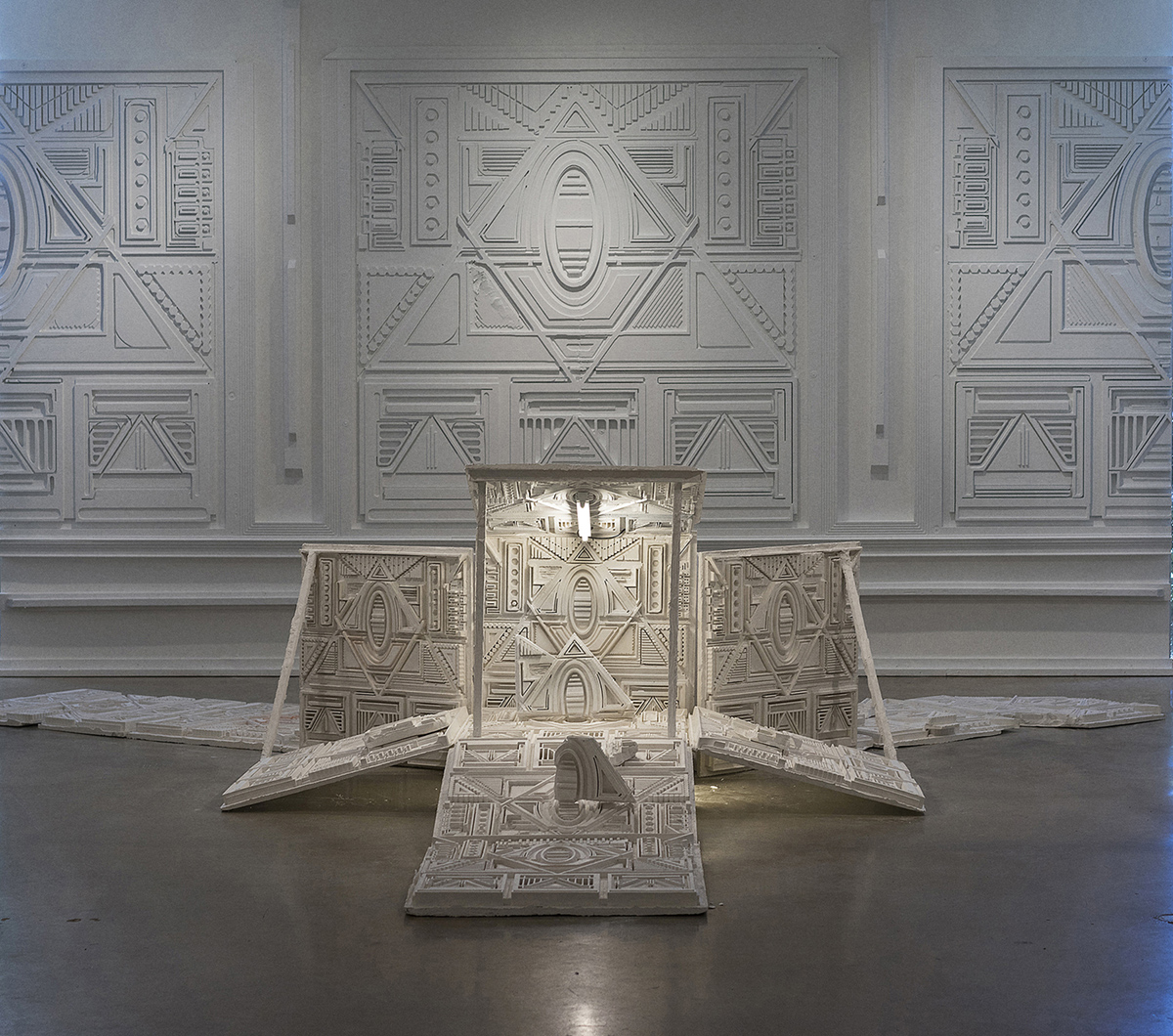 A white sculpture comprised of arranged flat panels with moldedgeometric patterns sits on the floor in a gallery space. The sculpture is situatedin front of a wall comprised of reliefs depicting the same patterns and textures on a larger scale. The sculpture is illuminated by a lightbulb, which increases the contrast of its patterning.