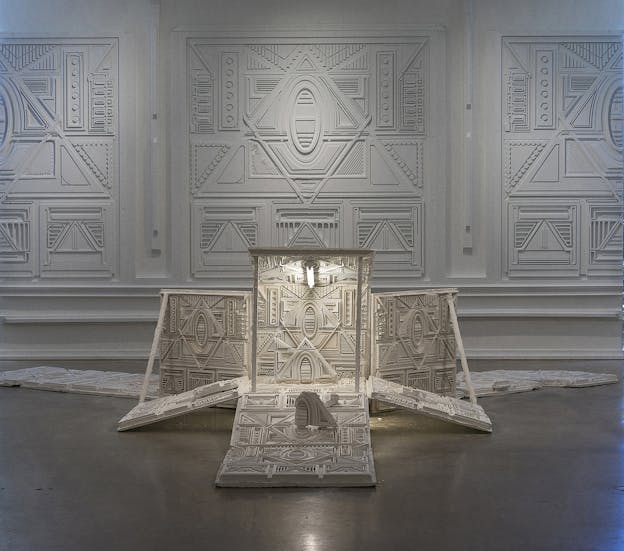 A white sculpture comprised of arranged flat panels with moldedgeometric patterns sits on the floor in a gallery space. The sculpture is situatedin front of a wall comprised of reliefs depicting the same patterns and textures on a larger scale. The sculpture is illuminated by a lightbulb, which increases the contrast of its patterning.