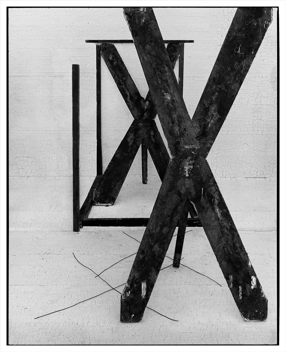 Two black Xs made from wooden beams overlap in the center of a black and white photograph. One is set further back than the other creating the illusion of space. On the floor pieces of wire mirror the shapes of the wooden beams.