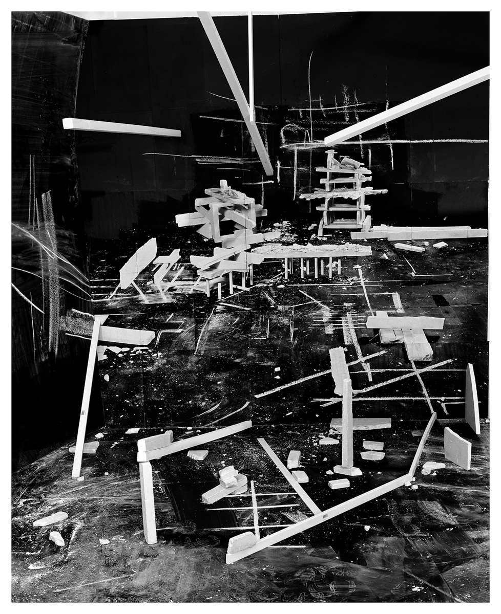 A high contrast black and white photograph of a dark space is filled with white rectangular forms and white chalk dust and lines, creating a sense of receding space and resembling a construction project in progress.