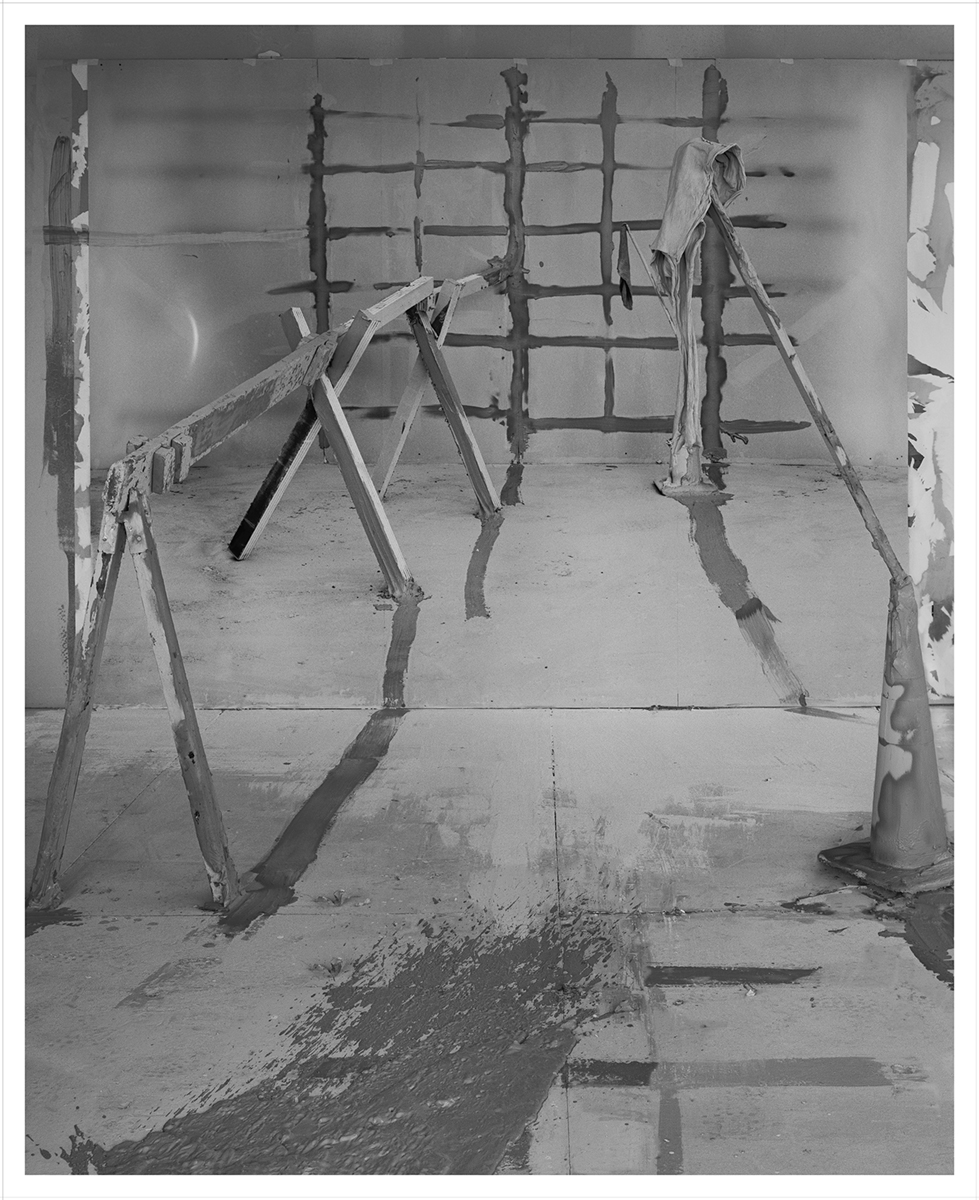 A black and white photograph of an industrial space containing  wooden barricades on each side. One of the barricades is held up by a traffic cone, and the barricades extend into a second room with a back wall painted to resemble a scaffolding’s shadow.