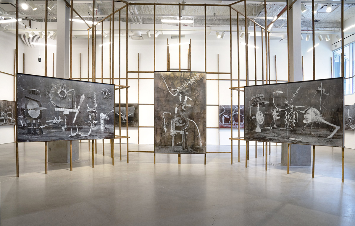A gallery installation of a network of intersecting wooden beams upon which three large-scale black and white photographs are mounted. The beams extend to the ceiling, across the room, and far back into the space. The textured photographs depict industrial scenes assembled by the artist using construction materials, such as two-by-fours, metal piping, and cinder blocks.