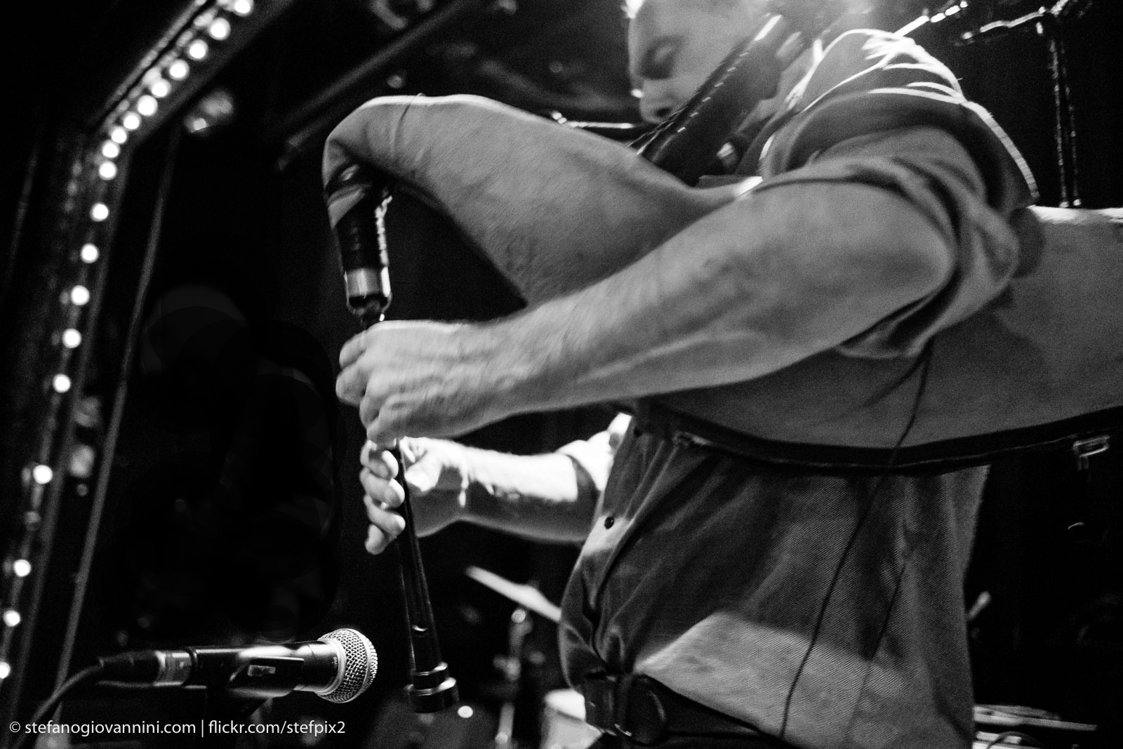 David Watson’s side profile as he plays the bagpipes on a stage in a concert venue. His eyes are closed and a microphone is pointed towards the bagpipes to amplify the sound. The photo is in black and white.