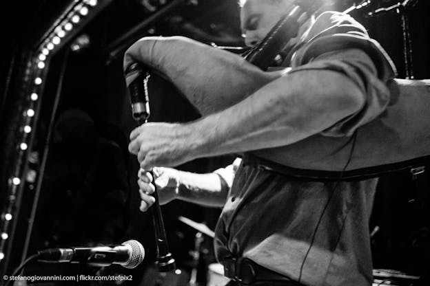David Watson’s side profile as he plays the bagpipes on a stage in a concert venue. His eyes are closed and a microphone is pointed towards the bagpipes to amplify the sound. The photo is in black and white.