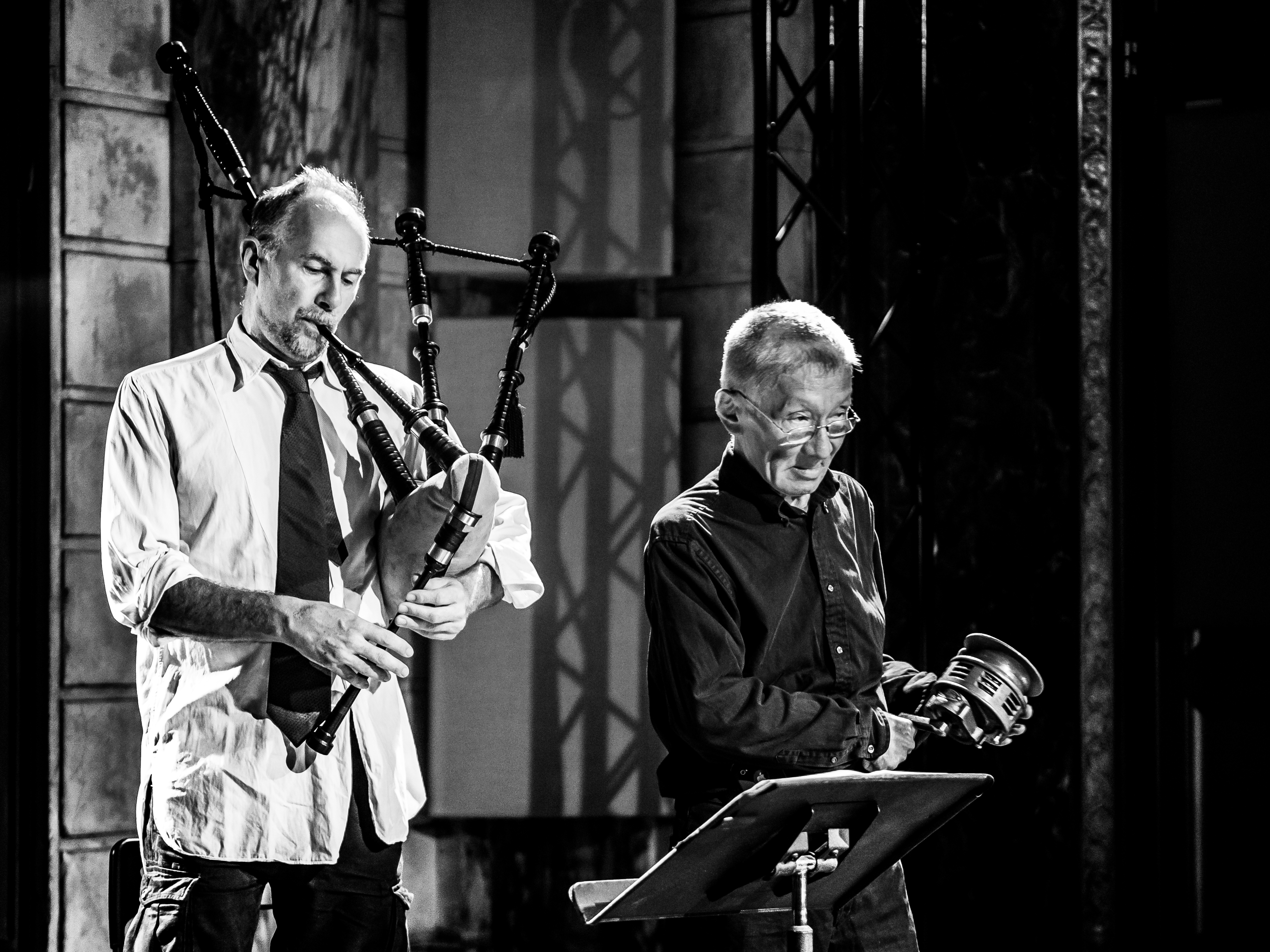 David Watson plays the bagpipes standing next to Yoshi Wada, who is playing a small handheld instrument on a stage in front of a music stand. Watson wears a light button up shirt with his sleeves rolled up and a black tie, and Wada wears a dark button up shirt and wire-framed glasses. The photo is in black and white.