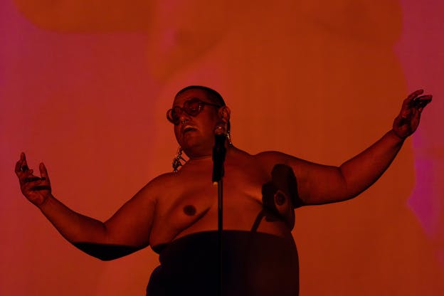 Dorian Wood stands behind a microphone with her arms out the sides and her head tilted, wearing heart-shaped sunglasses. She appears in front of a red, orange, and pink background and is bathed in orange light. Part of her body is in shadow and she does not wear a top.
