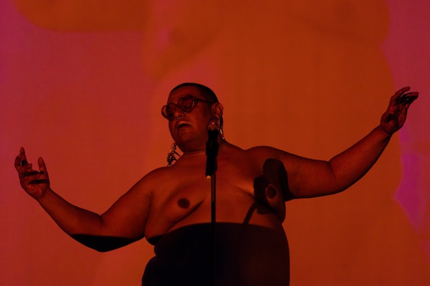 Dorian Wood stands behind a microphone with her arms out the sides and her head tilted, wearing heart-shaped sunglasses. She appears in front of a red, orange, and pink background and is bathed in orange light. Part of her body is in shadow and she does not wear a top.  