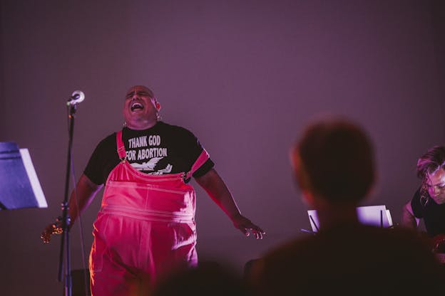 Dorian Wood sings on stage in front of a purple background behind a microphone. She wears pink overalls and a t-shirt that reads “THANK GOD FOR ABORTION” and next to her is a music stand. Towards the right of the image, a blurry silhouette of an audience member partially blocks a guitarist on the stage.