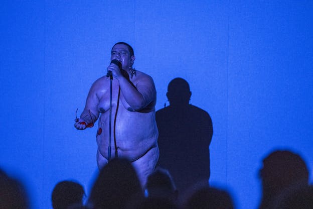 Dorian Wood stands naked on a stage singing in front of a blue background. She holds a microphone in one hand and a pair of heart-shaped sunglasses in the other and is illuminated by deep blue light. She is partially obscured by silhouettes of audience members in the foreground.