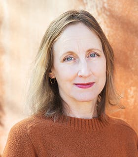 Peggy Piacenza smiles slightly at the camera with her body turned slightly to the side. She wears a burnt orange sweater, and hoop earrings against a brightly lit, warm toned wall.