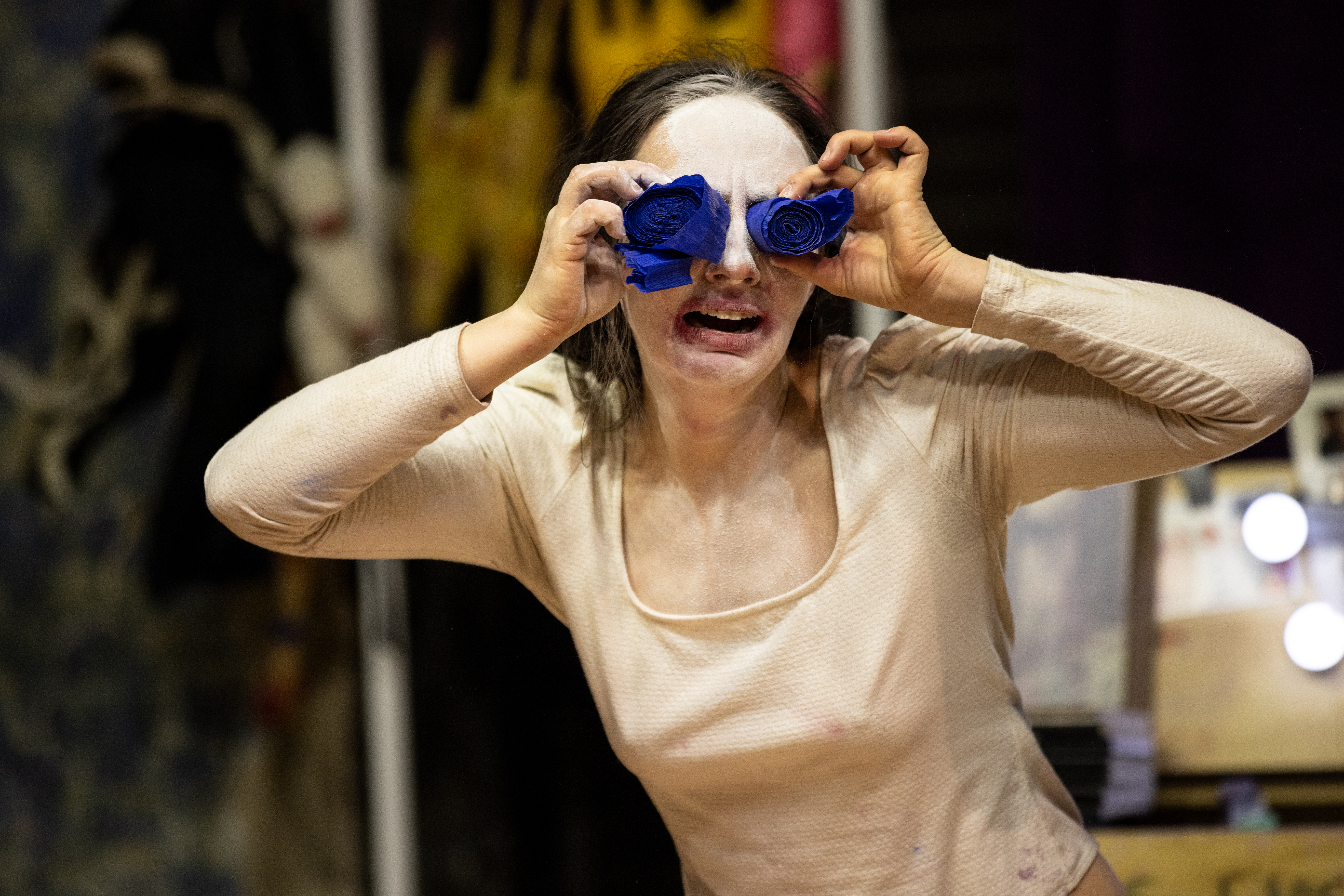 Alex Tatarsky, with their face painted white, presses two rolled-up blue streamers against their eyes while grimacing. They wear a beige long-sleeve top, and the background contains a cluttered performance area with various objects and a dressing table mirror with lights.