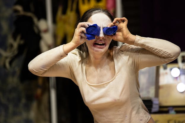 Alex Tatarsky, with their face painted white, presses two rolled-up blue streamers against their eyes while grimacing. They wear a beige long-sleeve top, and the background contains a cluttered performance area with various objects and a dressing table mirror with lights.