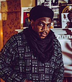 Jamal R. Moore gazes directly at the camera with his body turned slightly to the side. He wears a black hat, scarf, and pattered top. A fireplace mantle with and yellow wallpaper are visible in the background.