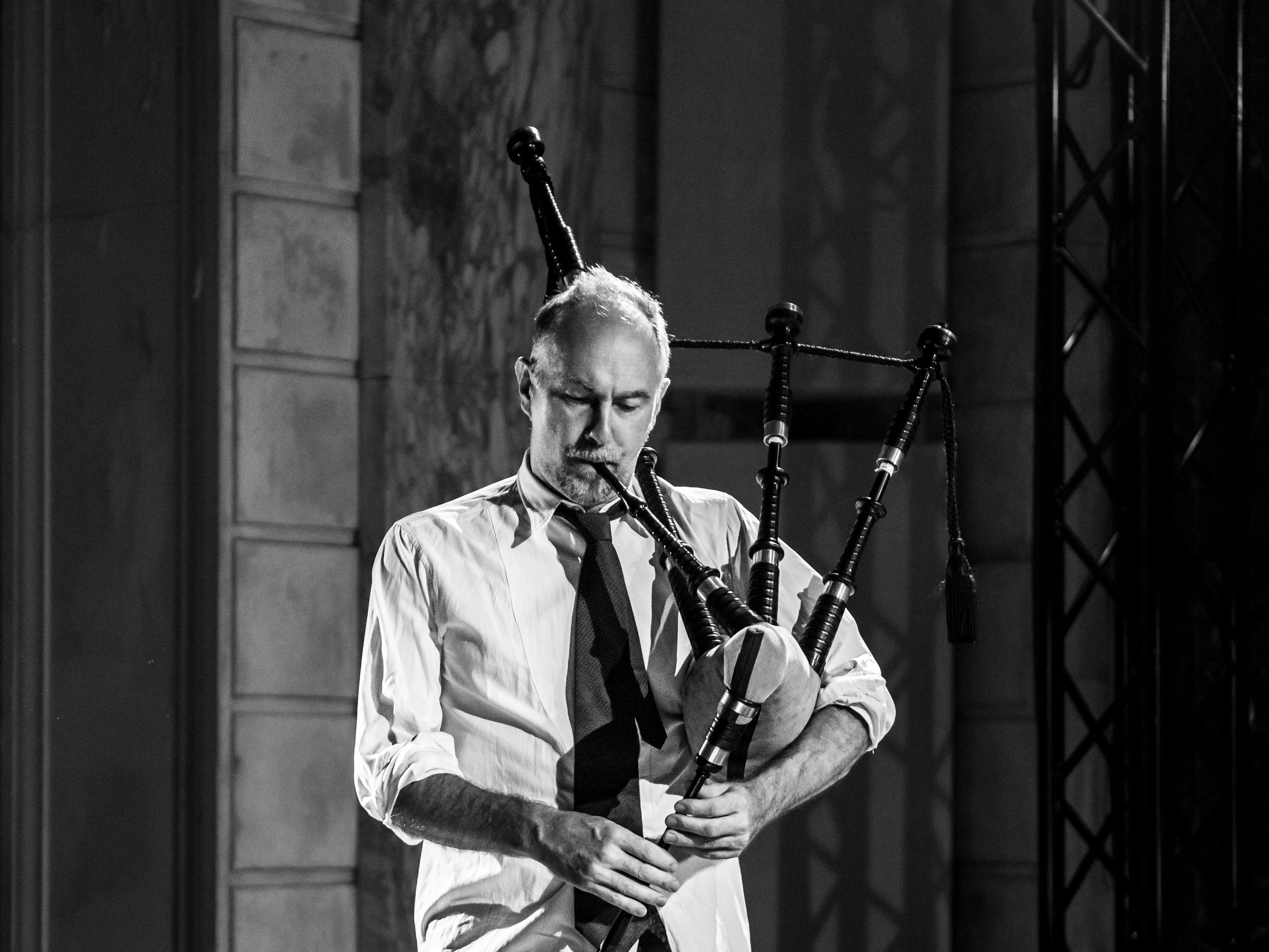 A black and white photo of David Watson playing the bagpipes. He wears a light button up shirt with his sleeves rolled up and a black tie.