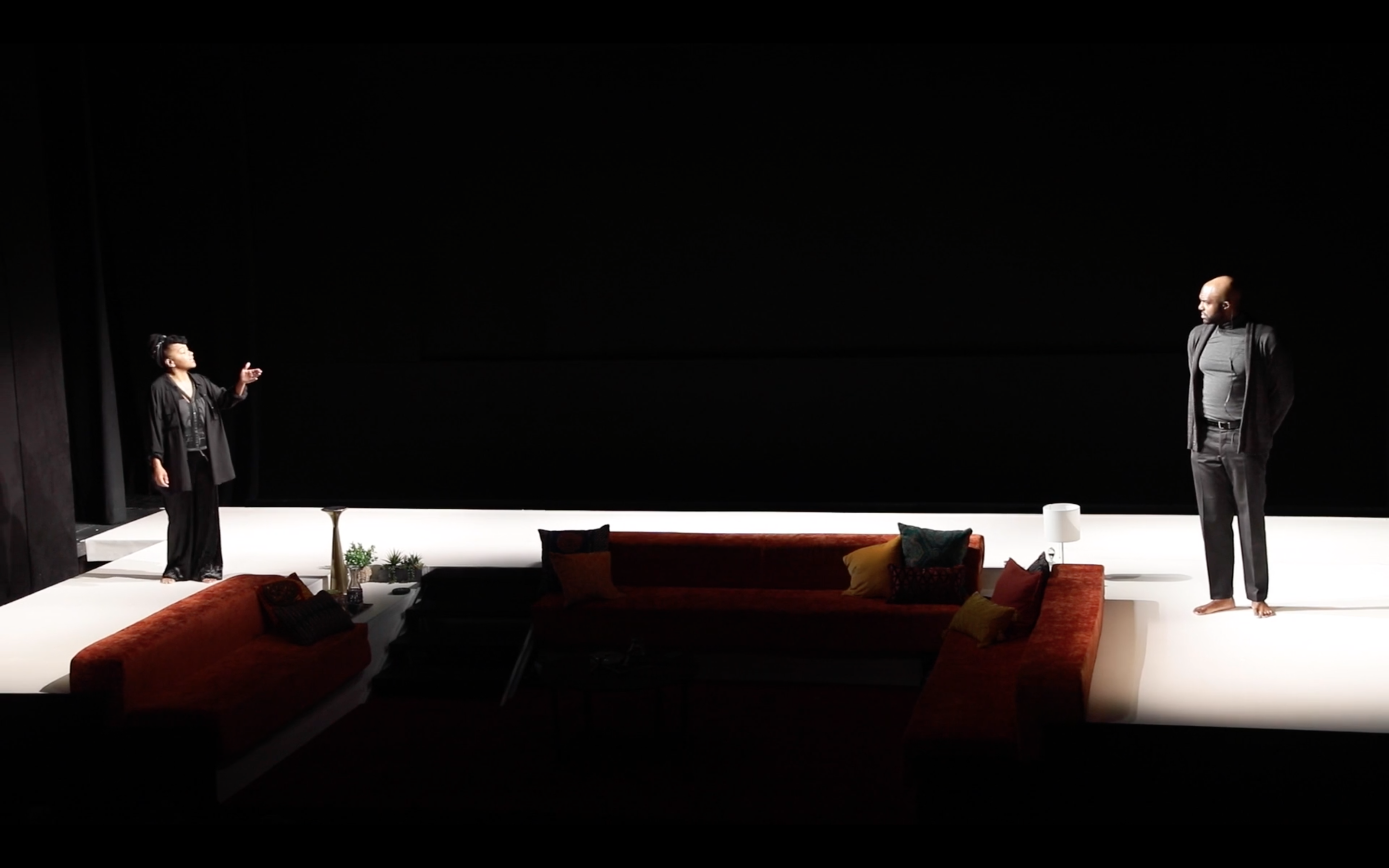 Crystal Dickinson (left) and Carl Clemons-Hopkins (right) perform on a stage. Dickinson, dressed in black, stands on an elevated white platform, gesturing with one hand. Clemons-Hopkins, wearing a dark suit and turtleneck, stands barefoot at a distance with hands behind his back. The set includes an orange sectional sofa with colorful pillows, a small table, and a white lamp. The background is dark, with bright lighting on the platform and performers.