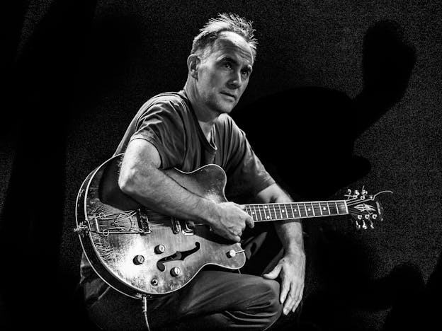 A black and white photo of  David Watson sits with a hollow-body electric guitar against a dark background. He wears a short-sleeved shirt, resting one arm on the guitar while the other hand is placed on his knee.