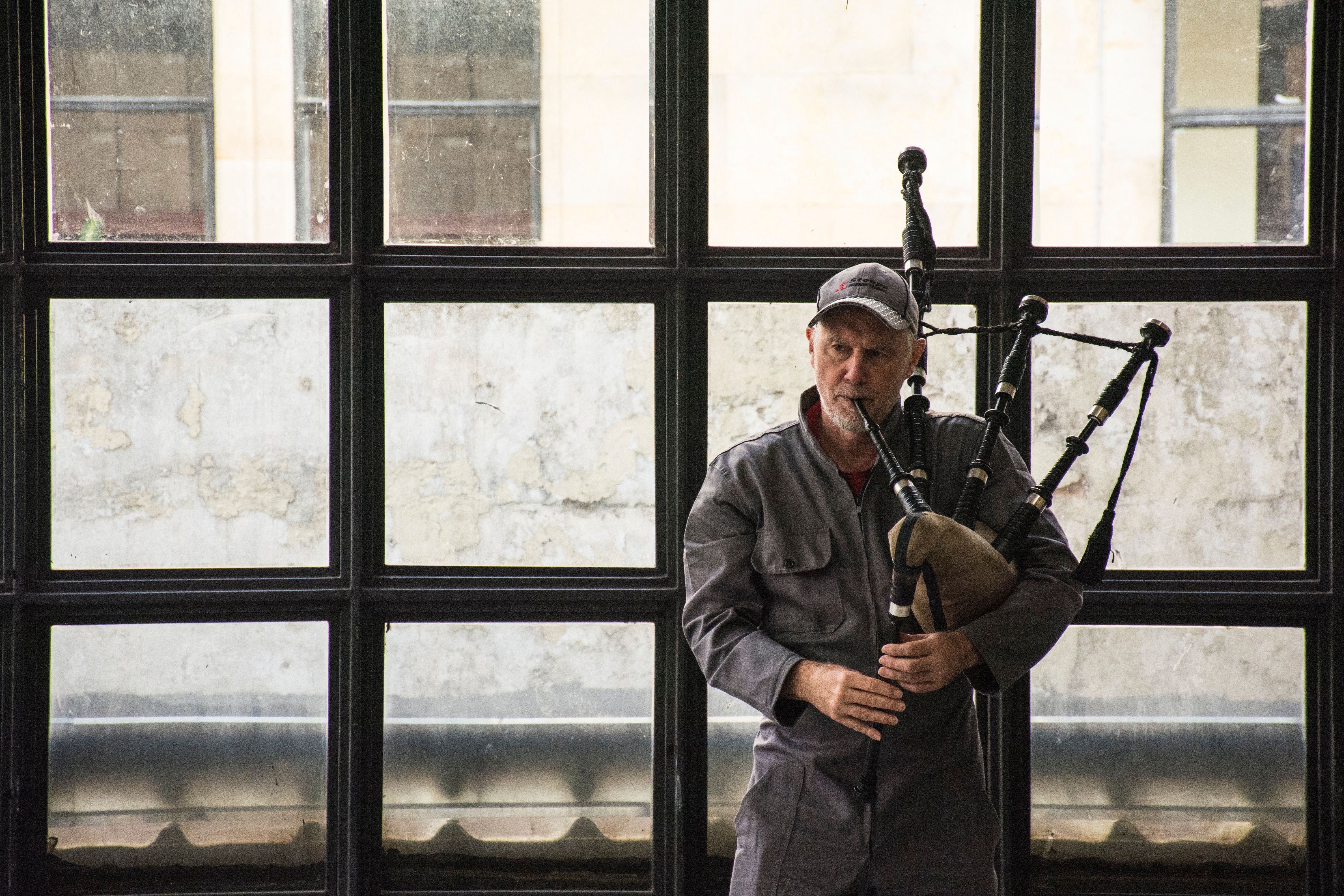 David Watson, wearing a gray work jumpsuit and a baseball cap, plays the bagpipes indoors. He stands in front of a large industrial-style window with a weathered wall visible in the background, holding the bagpipes under his arm.
