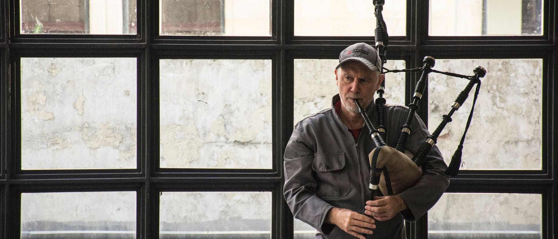 David Watson, wearing a gray work jumpsuit and a baseball cap, plays the bagpipes indoors. He stands in front of a large industrial-style window with a weathered wall visible in the background, holding the bagpipes under his arm.