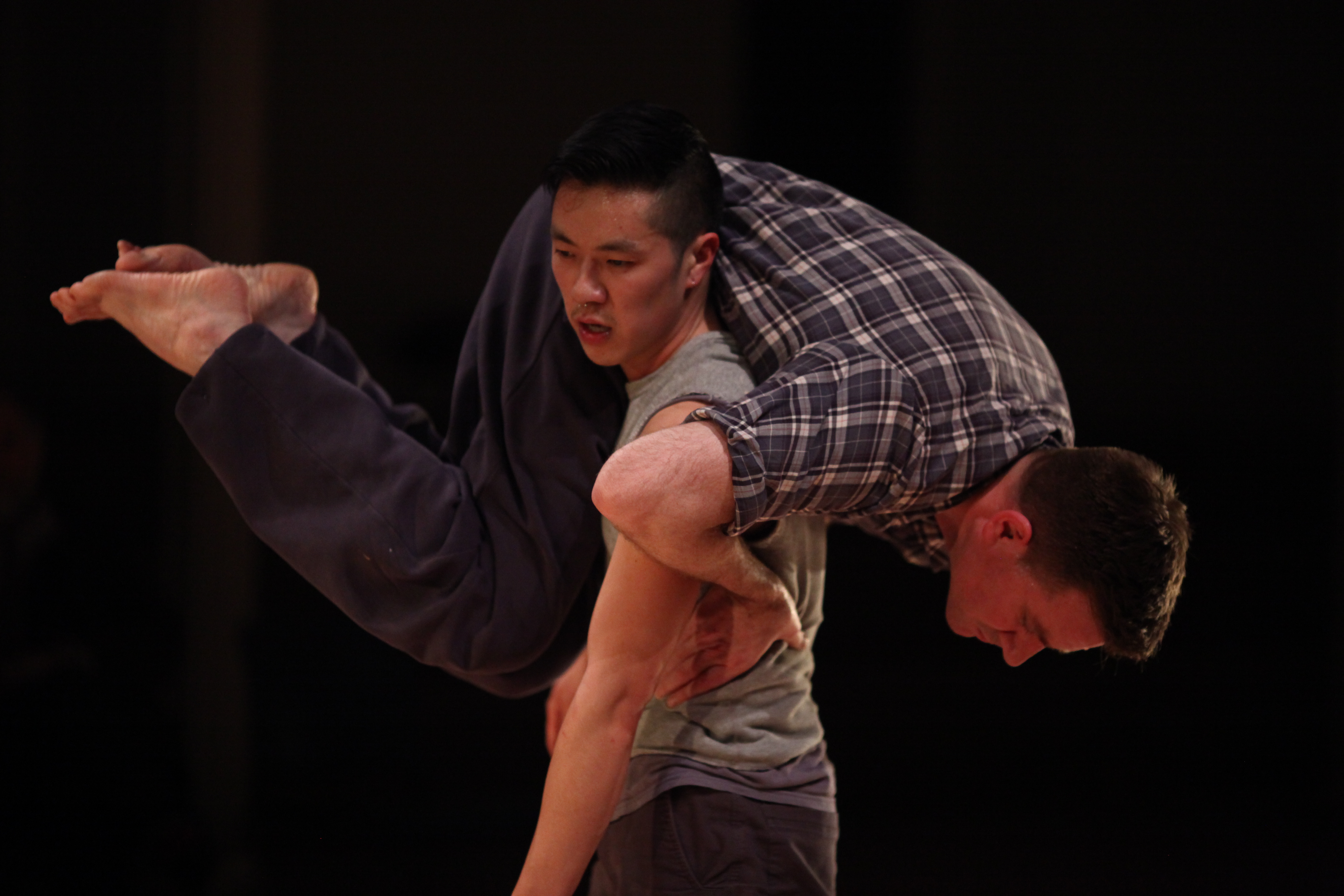 Enrico D. Wey, wearing a gray sleeveless shirt and dark pants, supports Jon Kinzel, who is draped over his back wearing a plaid long-sleeve shirt and dark pants. Their bodies are intertwined in a movement against a dark background.