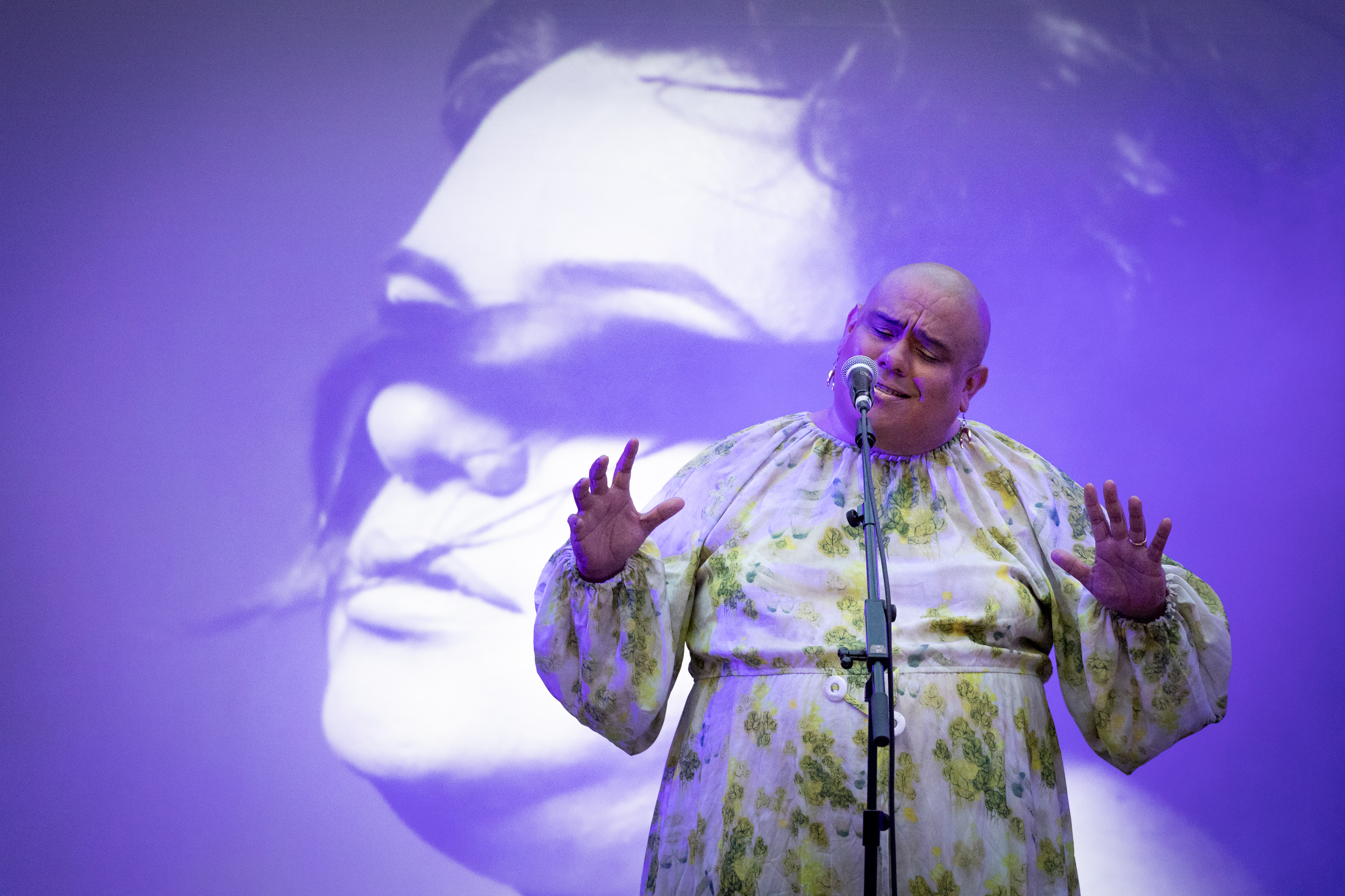 Dorian Wood stands at a microphone in front of a projected purple-and-white image of a person with a long lock of hair covering their eyes. She wears a green and white patterned long sleeved dress and is illuminated by soft purple light. 