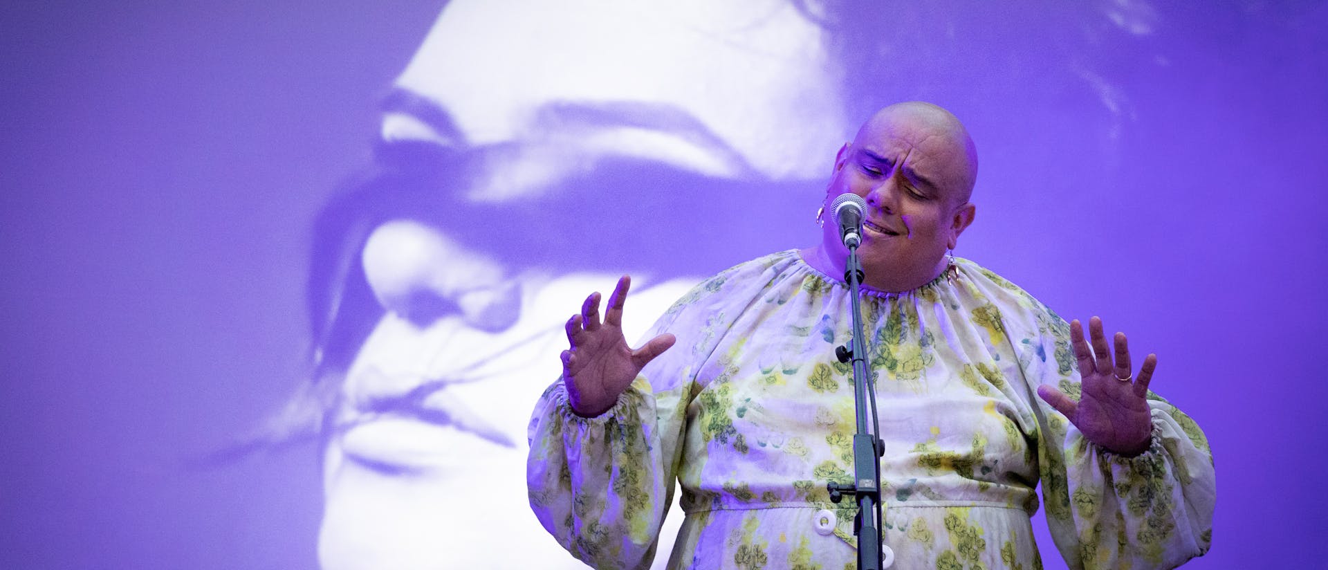 Dorian Wood stands at a microphone in front of a projected purple-and-white image of a person with a long lock of hair covering their eyes. She wears a green and white patterned long sleeved dress and is illuminated by soft purple light.