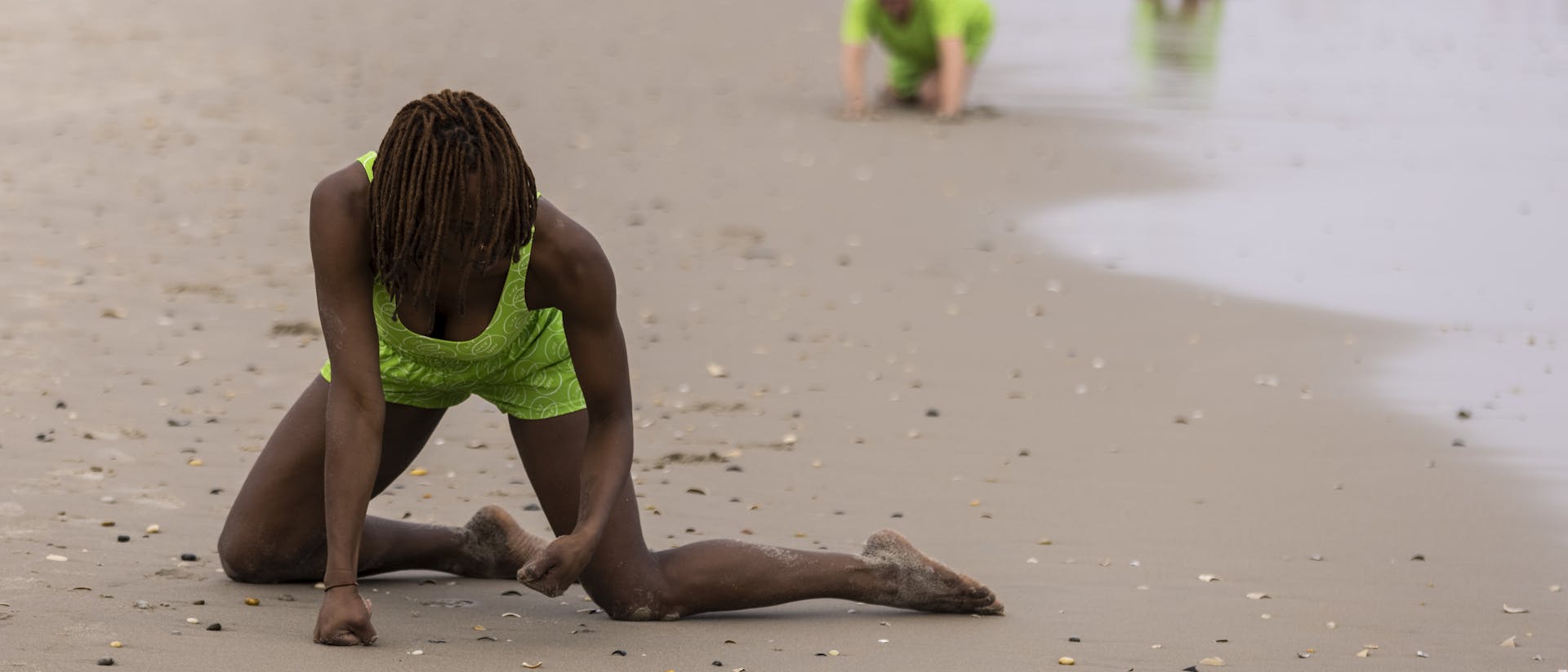Shayla-Vie Jenkins kneels on a beach in a neon green outfit, pressing her fists into the ground as she extends her legs sideways. The sky is overcast and, in the background, additional performers dressed in similar neon green outfits crawl across the sand. Beachgoers are seen in the distance walking along the shoreline.
