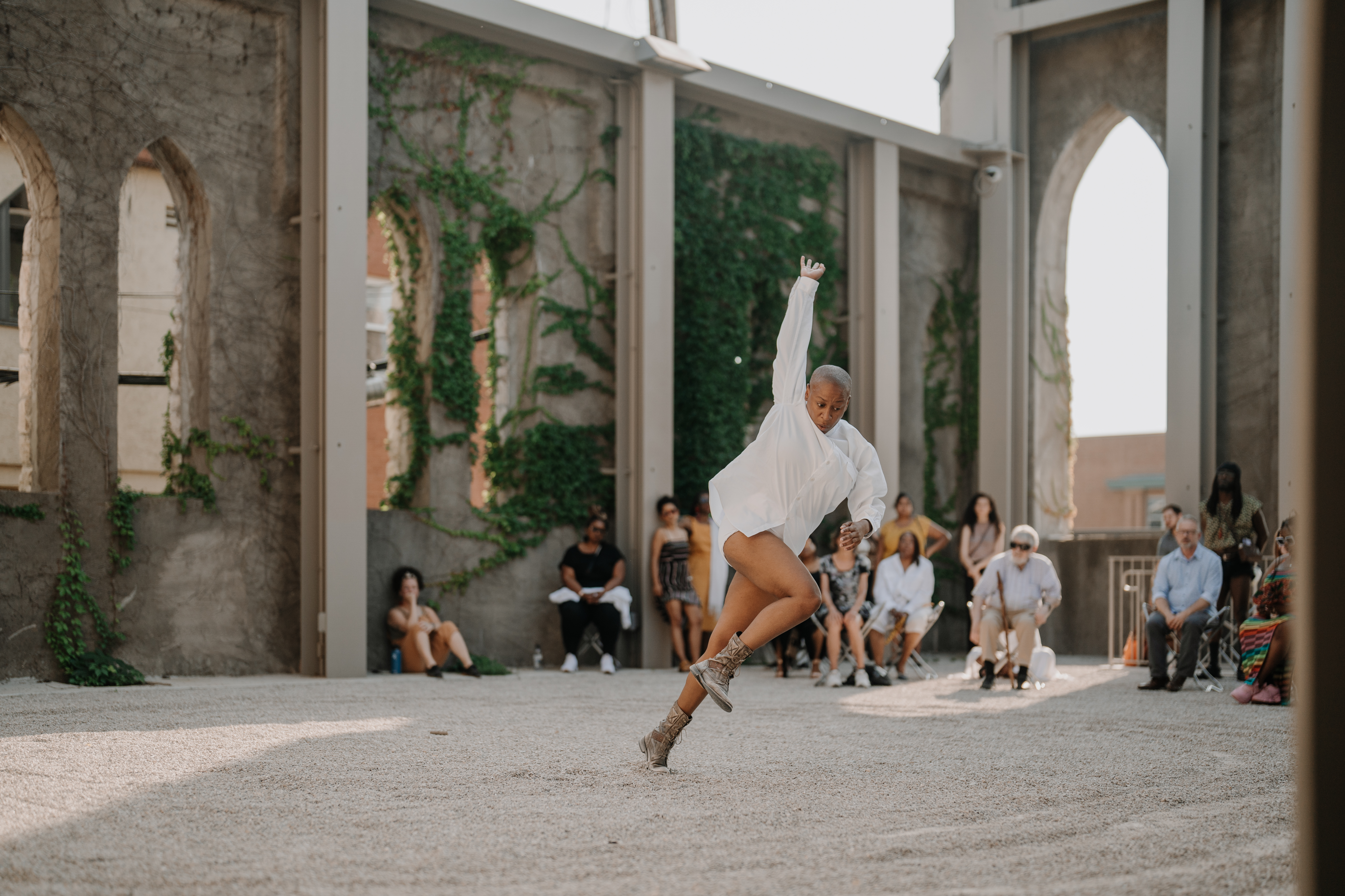 Jennifer Harge performs a dynamic movement in an outdoor space with tall, ivy-covered walls and arched windows. She wears a white long-sleeved garment and lace-up boots, mid-step with one arm extended upward and her body leaning forward. In the background, an audience watches attentively in the sunlit venue with gravel flooring.