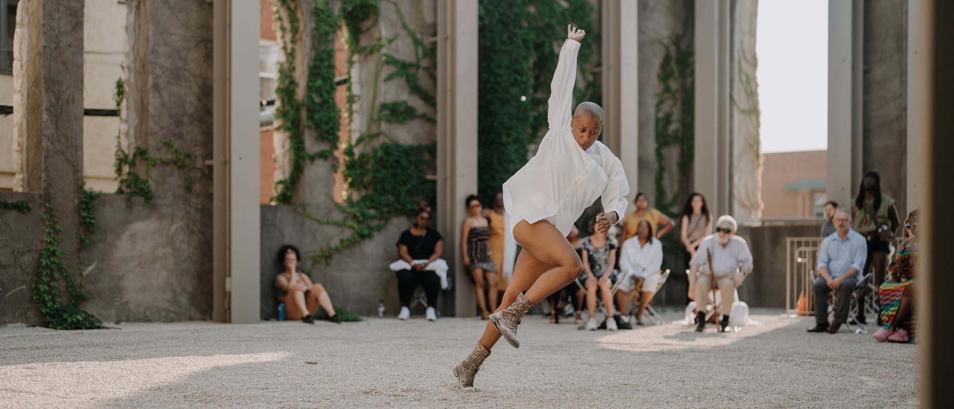 Jennifer Harge performs a dynamic movement in an outdoor space with tall, ivy-covered walls and arched windows. She wears a white long-sleeved garment and lace-up boots, mid-step with one arm extended upward and her body leaning forward. In the background, an audience watches attentively in the sunlit venue with gravel flooring.