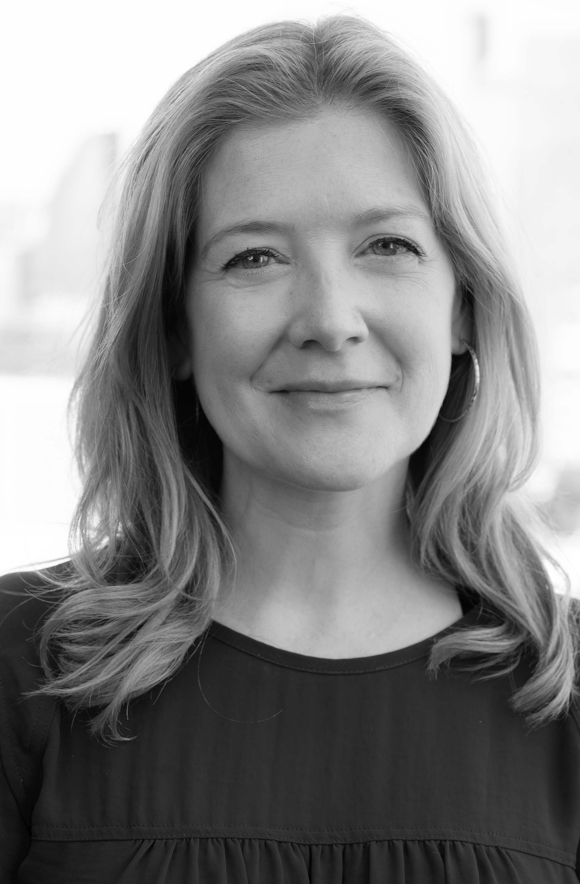 Black and white portrait of Heather Bhandari looking towards the camera with shoulder length blond hair, wearing a black crew neck shirt and hoop earrings.