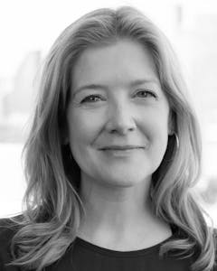 Black and white portrait of Heather Bhandari looking towards the camera with shoulder length blond hair, wearing a black crew neck shirt and hoop earrings.