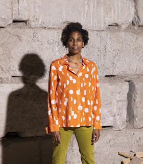 Photograph of Karyn Olivier looking towards the camera in front of a stone wall. She wears a colorful necklace, an orange button down shirt with white polka dots, and light green pans. Her shadow is visible in the framer.