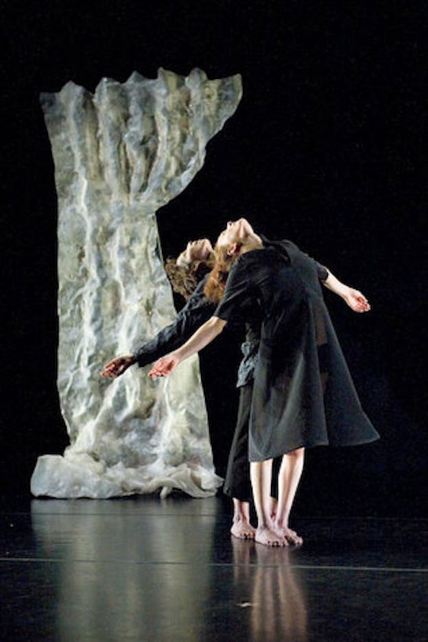 Two performers dressed in black outfits stand behind each other against a black backdrop. Both of them raise their heads up while spreading their arms out. A piece of white ruffle fabric tinged with washed green and blue sits vertically in the background. 
