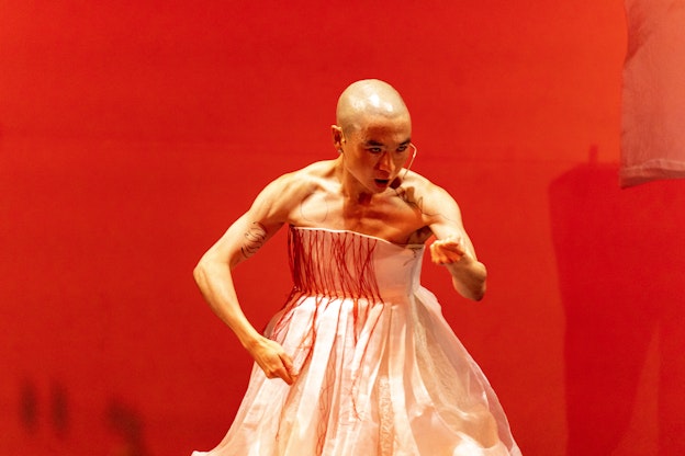 eddy stares intently toward the right side of the image, her head bowed slightly. Her left hand is clenched into a fist in front of her, her right elbow bent slightly to the side, the muscles in her shoulders and arms tensed. Her head is shaved and she is wearing a white dress with red threads hanging down the front.