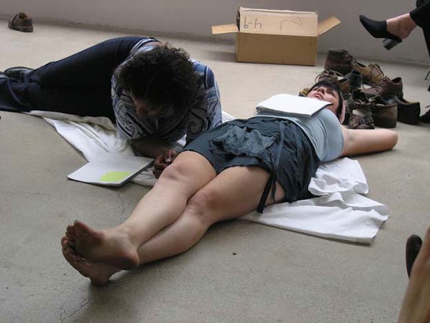 Two people are on top of a folded white fabric, one in a reclining position and the other one lying on their back. Behind them are pairs of brown leather laced shoes and a cardboard box.