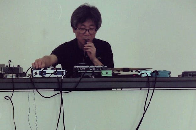 A person dressed in a black t-shirt and glasses, sits behind a desk that is occupied by tangled cables.