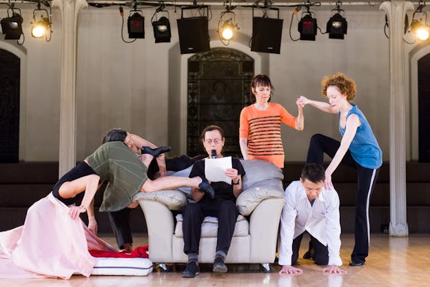 Rainer sitting on a couch reading from a sheet of paper while performers around her perform various motions.