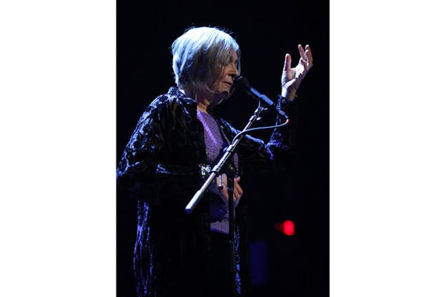 Photograph of a person with chin-length gray hair, a purple shirt and dark cardigan in front of a microphone performing with closed eyes.  