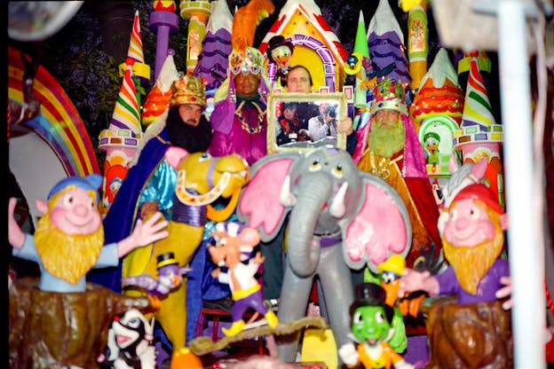 There is a play castle within a dark space. The steeples of the castle are covered in rainbow stripes and bright lights. In the middle of this castle, large plastic toys are arranged. There is an elephant, gnomes, and a camel along with numerous other smaller toys. Behind these toys, four people stand. Three of these people wear play royalty costumes in fushia and purple. A fourth person stands and holds a gold framed image of someone taking a photograph in a crowd. 