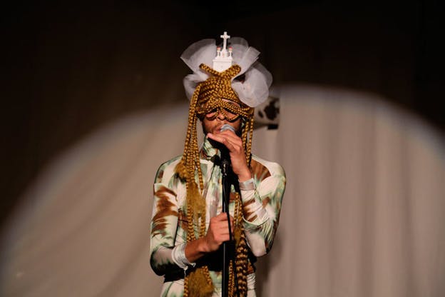 Performer holding a microphone, illuminated by a spotlight, their eyes covered in long, honey-colored braids and on the top of their head a white church ornament and white tulle. 