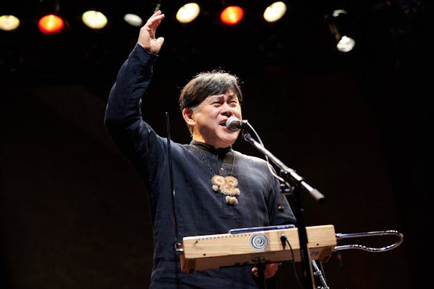 Koichi Makigami performs on a brightly lit stage, he is facing the camera and stands behind a theremin and microphone. He looks towards the right with one hand raised by his head as he vocalizes into the microphone. He is wearing a dark henley shirt and a tan medallion necklace.