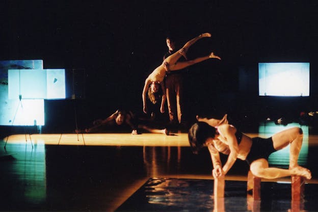 Three performers are in a dark space with blue hued projections in the background. In the foreground, a performer wearing a black top and black shorts balances their body on bricks on top of a pool of water. In the background another performer holds someone upside down.