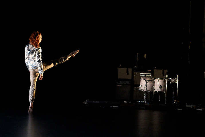 Performer faces away and extends one leg forward in relevé on the left side of a black-lit stage with a silver drum set in the right corner.