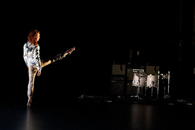 Performer faces away and extends one leg forward in relevé on the left side of a black-lit stage with a silver drum set in the right corner.