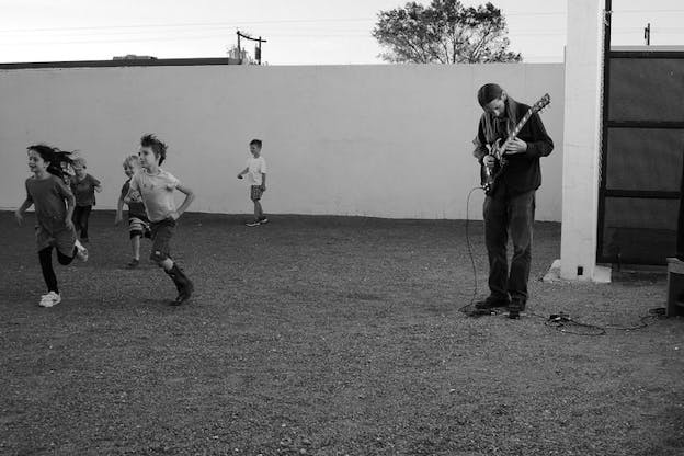 Black and white photograph of Barr in a yard with a low white wall playing the electrical guitar beside children running and smiling. 
