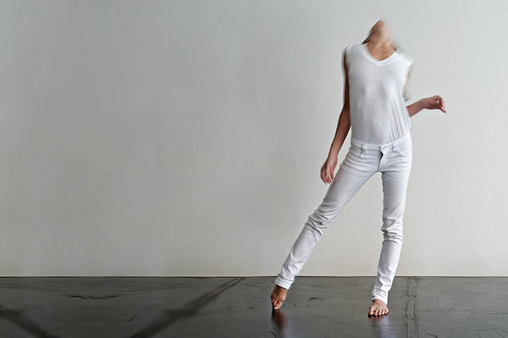 A blurred performance still of Maria Hassabi wearing all white and standing in front of a white wall. She puts her weight on her left leg as her right leg extends to the side. Her left arm is bent at the elbow and is close to her ribcage while her right arm is extended out. Her head is throw back and her chin points towards the ceiling. 