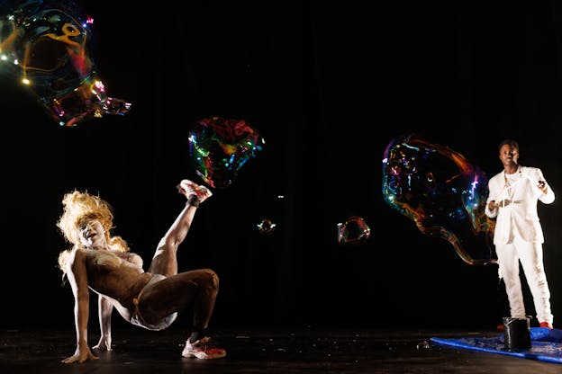 Crackhead Barney and Nile Harris perform on a dark stage lit from the sides with giant iridescent bubbles behind them. Crackhead Barney poses on the left side of the stage, she balances on her hands and right leg, belly up, with her left leg kicking upwards. Her face and body are smeared with white body paint, she wears a blond, curly wig, diaper-like underwear, tape covering her breasts, and red sneakers. Nile Harris stands on the right side of the stage in front of a paint bucket, he is making a bubble with a wand created from two sticks and a string. He is wearing a white suit and red dress shoes. 