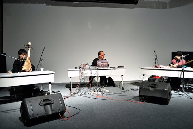 Three performers sit at three white tables in a white space/ On the left, a performer sits playing a mandolin. In the center, Stone sits at a desk in front of an open latop. Another performers plays an instrument at their desk on the right.